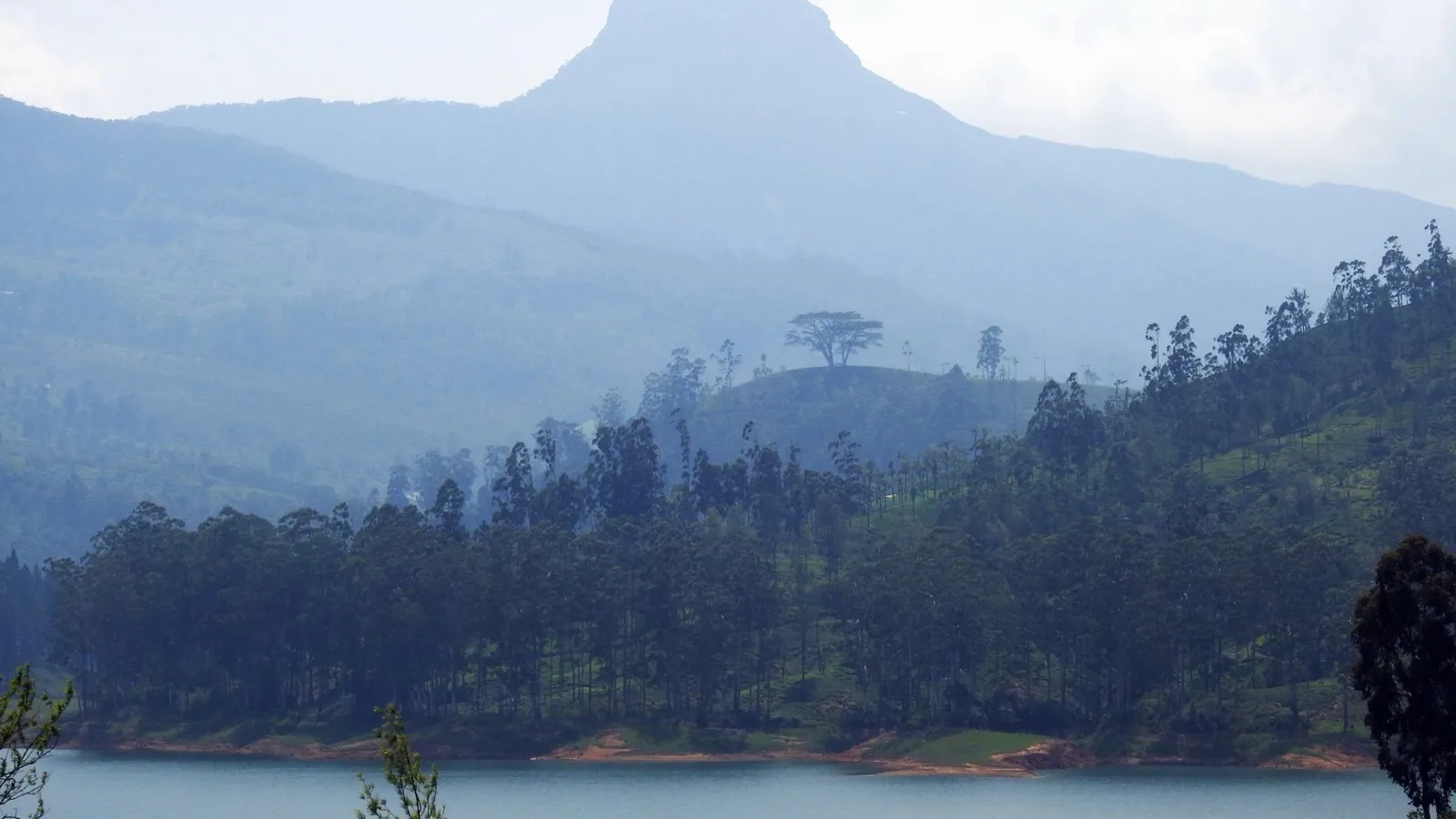 The pointed pyramid of Adam's Peak rising above forested hills and the still water of Maskeliya Reservoir