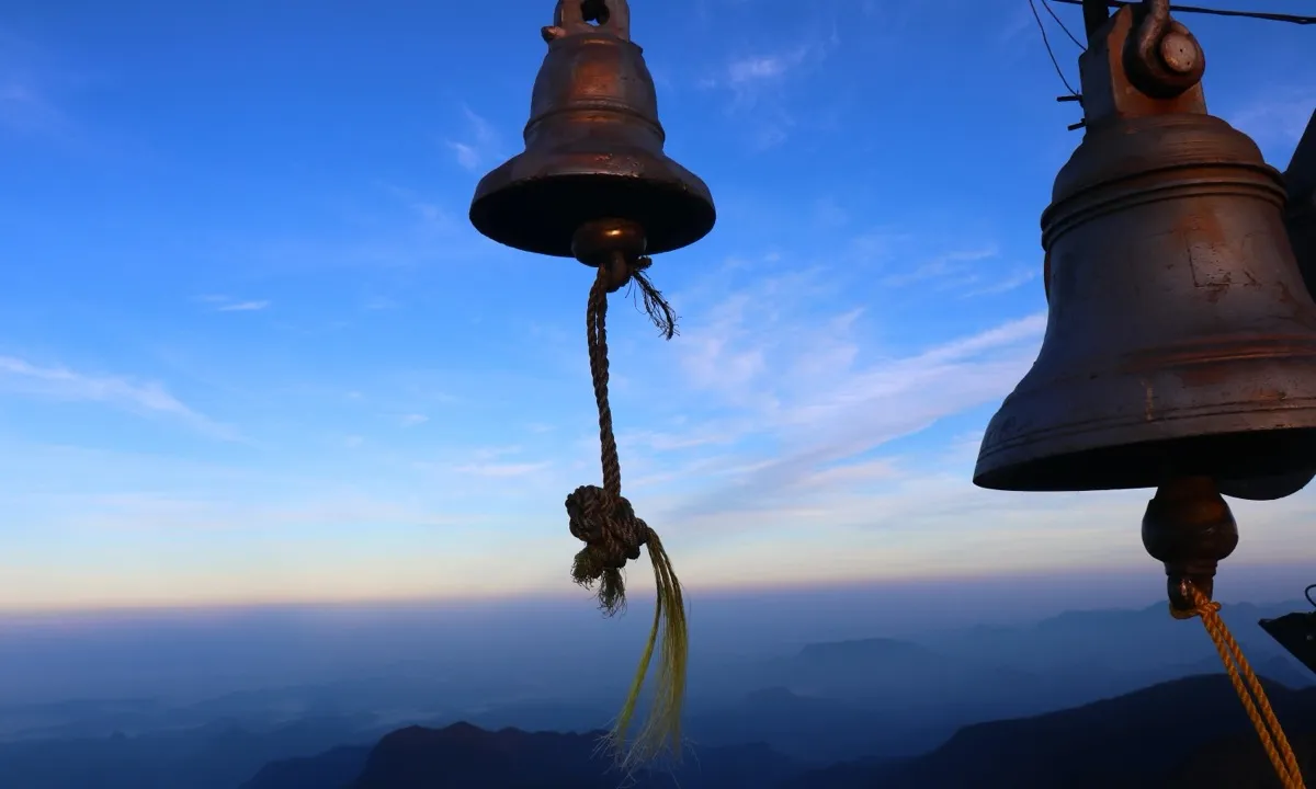 Two bronze bells with knotted rope pulls at the summit of Adam's Peak against a blue dawn sky over distant ridges