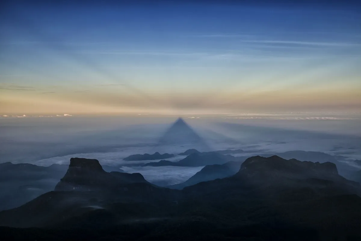 The pointed shadow of Adam's Peak projected onto a sea of cloud at sunrise, with dark ridges in the foreground
