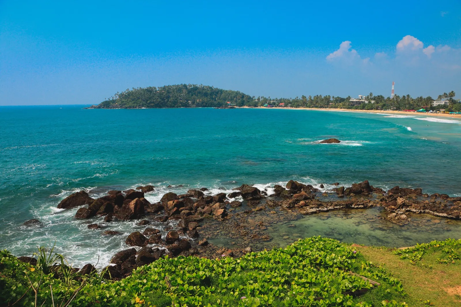 Mirissa bay on Sri Lanka's south coast under a clear blue sky, with turquoise water and dark rocks
