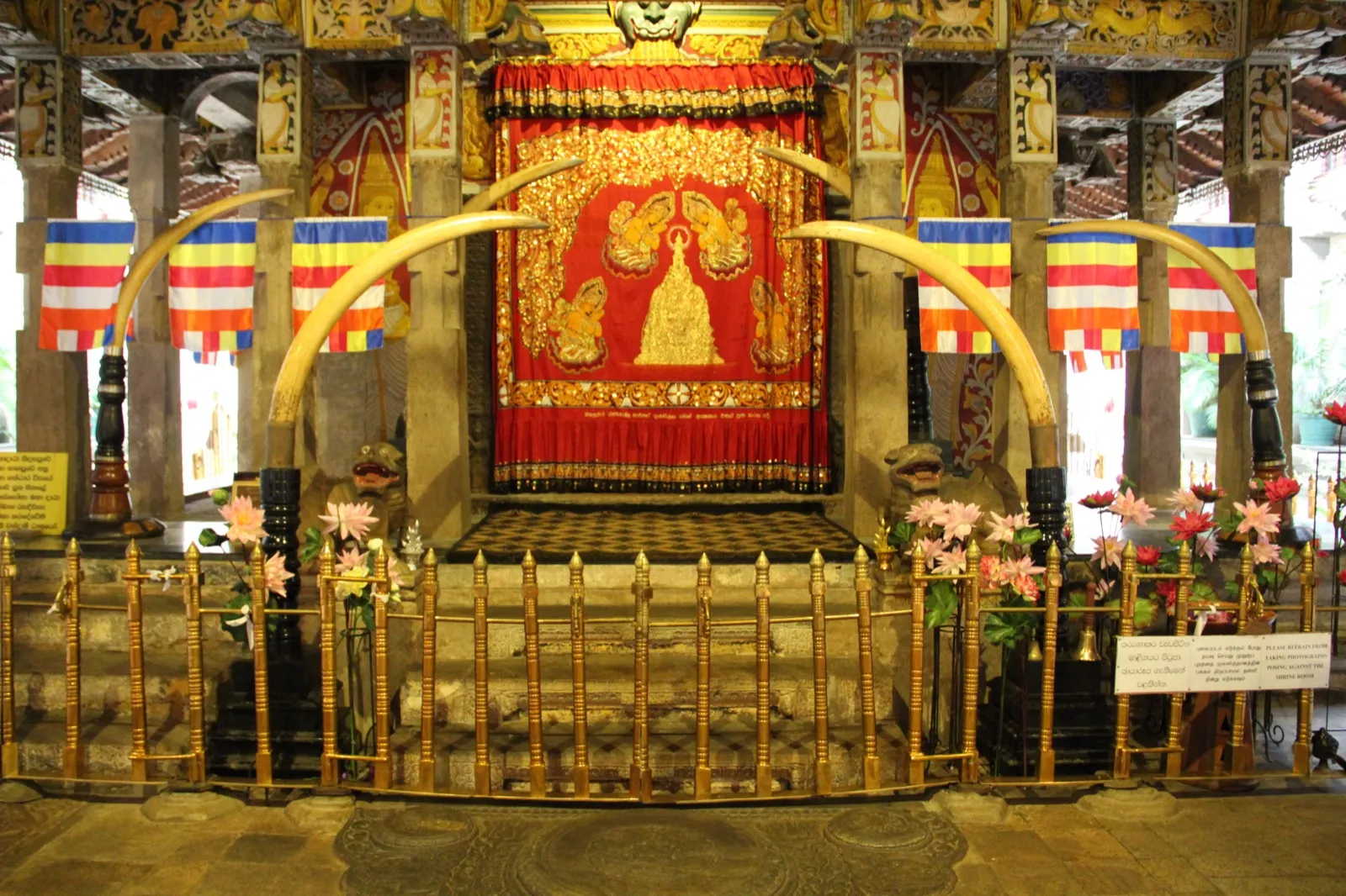 The ornate red-curtained shrine room door at the Temple of the Tooth in Kandy, framed by tusks and Buddhist flags, with a small sign asking visitors not to pose for photos