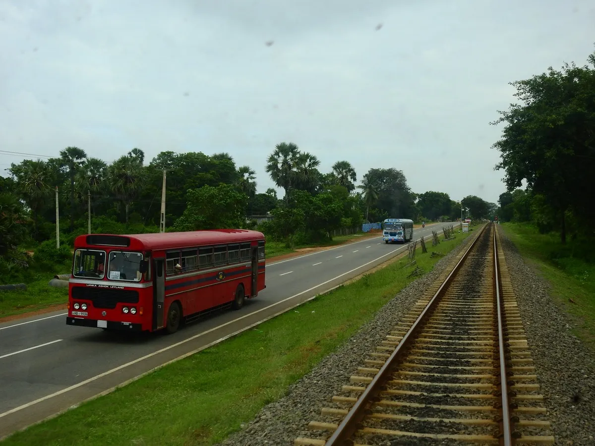 A red public bus and a blue private bus on a straight road running beside a railway track
