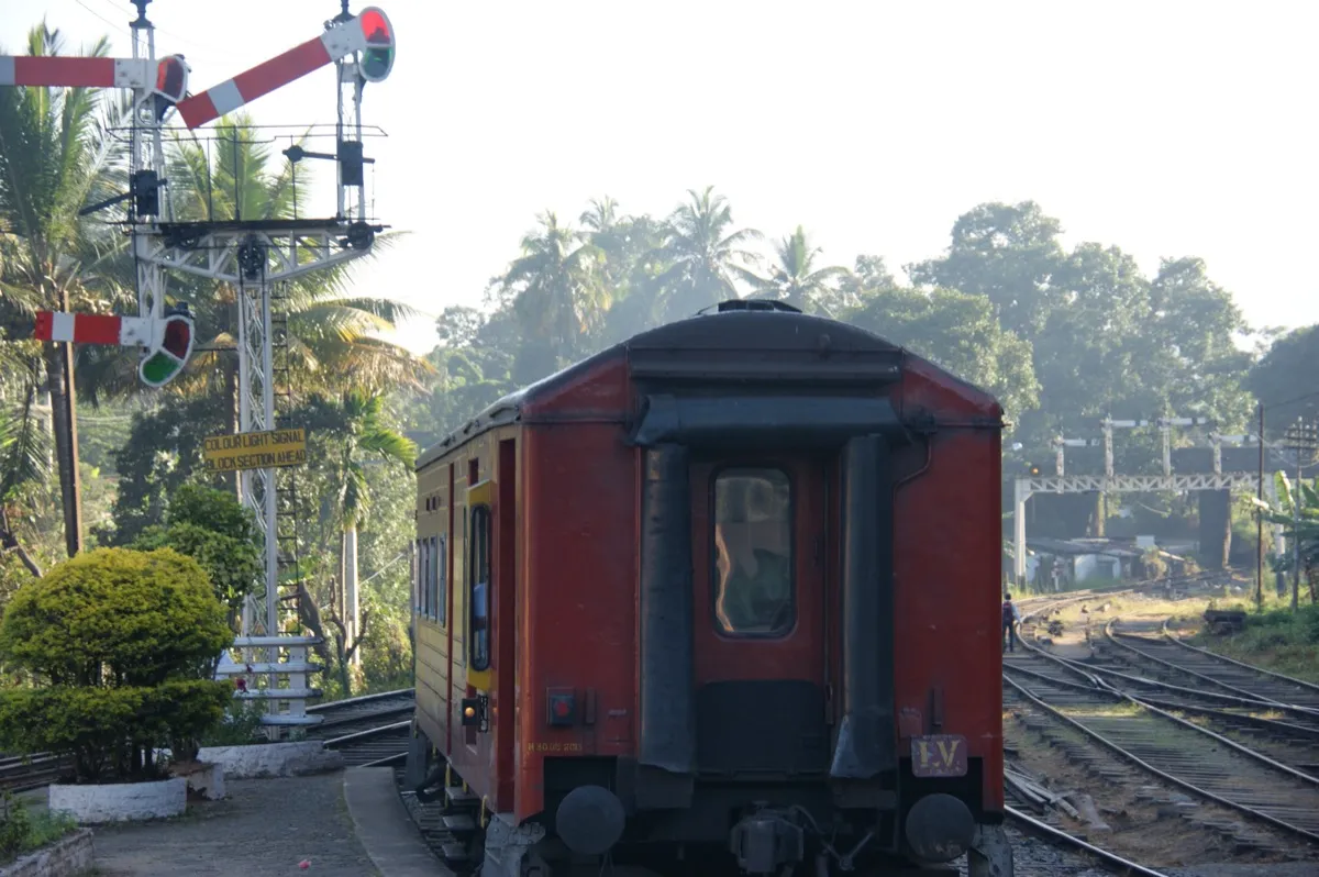 The end of a red Sri Lanka Railways carriage standing at Peradeniya Junction, with old semaphore signals beside the line and palms behind