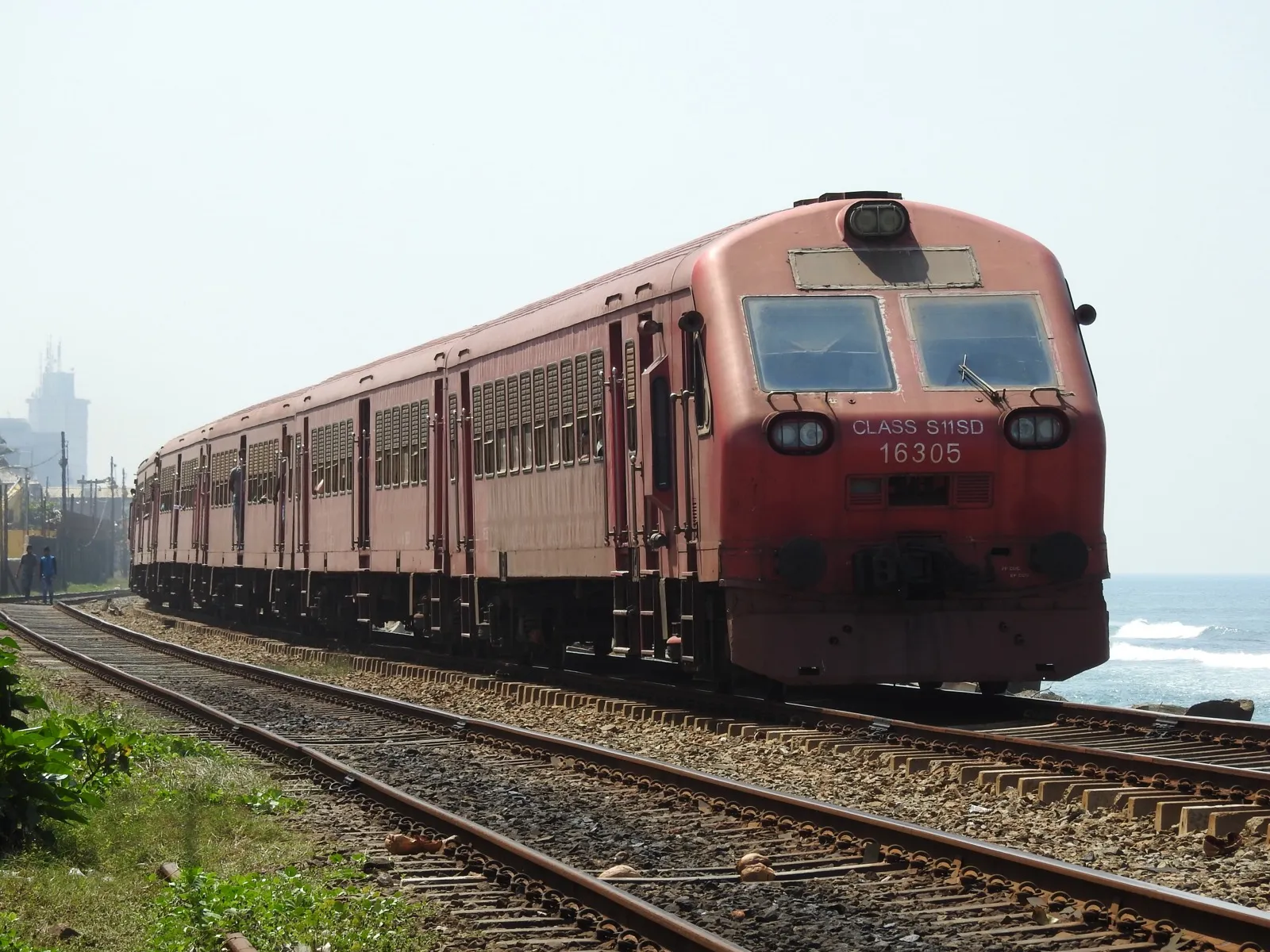 A red passenger train running on the seaside railway line in Colombo, with waves breaking just beside the track