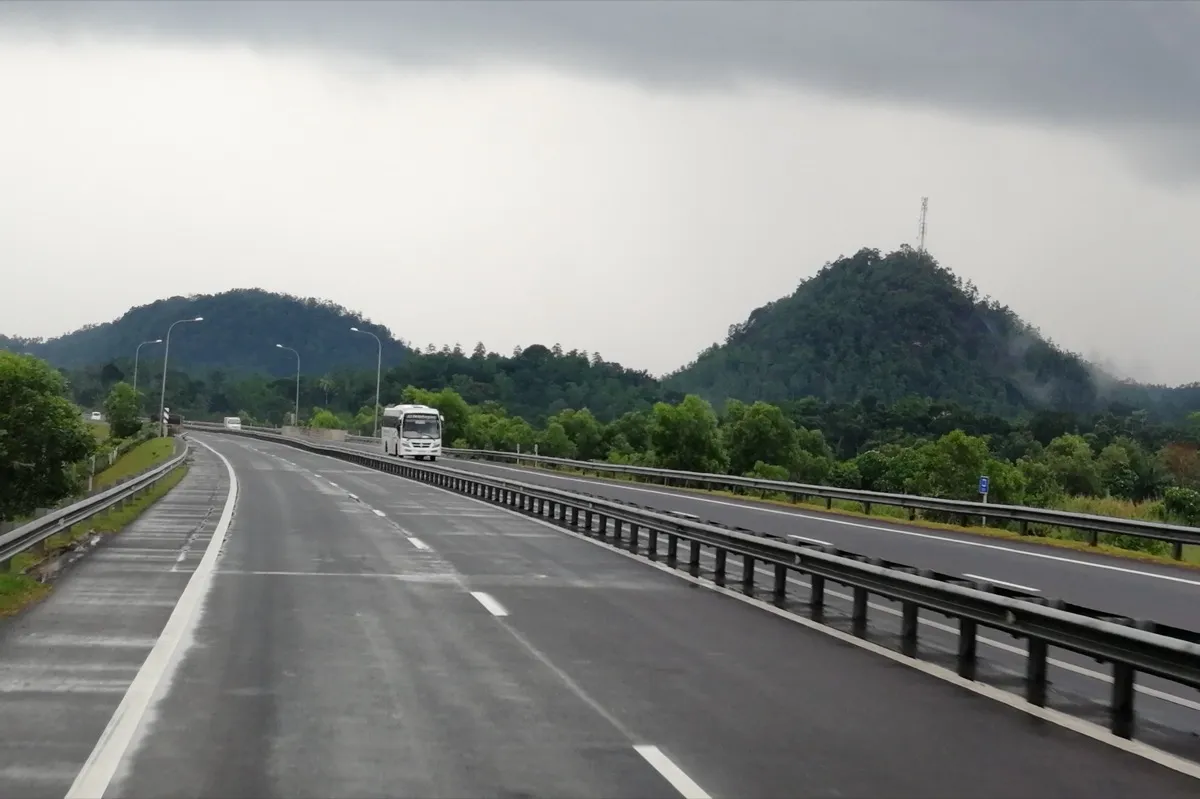 A wide expressway with guard rails running between green hills, with a white coach coming the other way