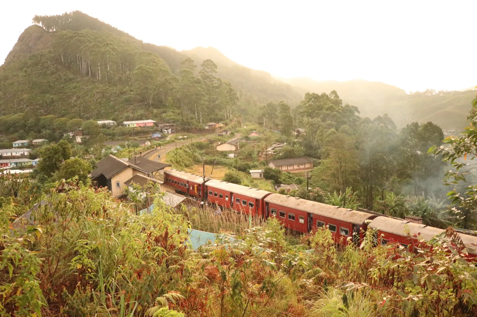 A red passenger train curving past estate houses through misty, forested hill country in Sri Lanka