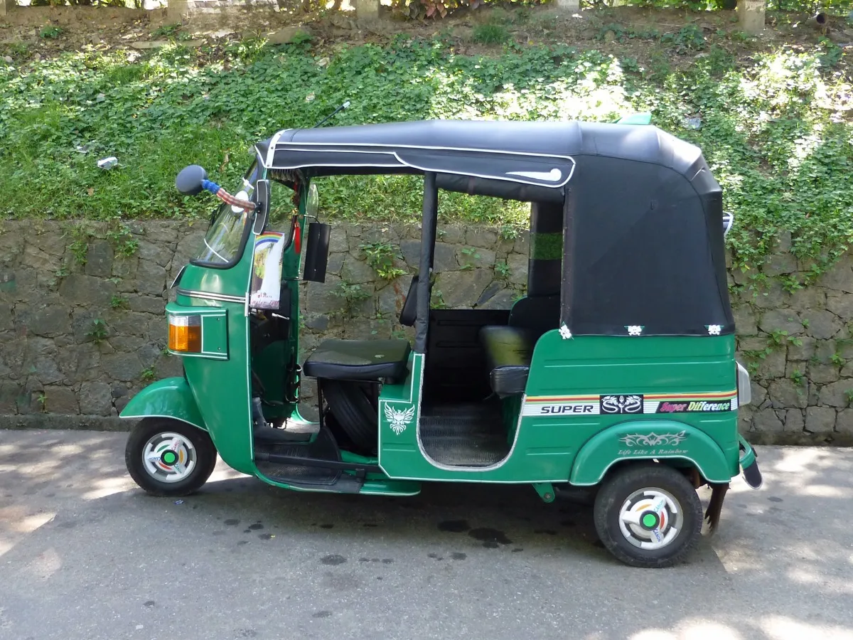 A green tuk-tuk with a black canopy parked beside a mossy stone wall on a street in Kandy