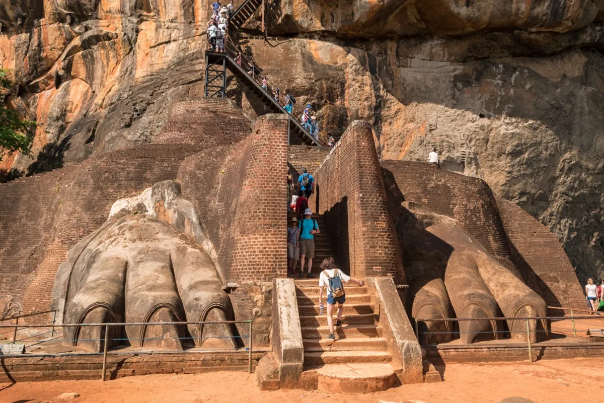 Two giant stone lion paws flank a brick stairway at Sigiriya, with metal stairs climbing the rock above