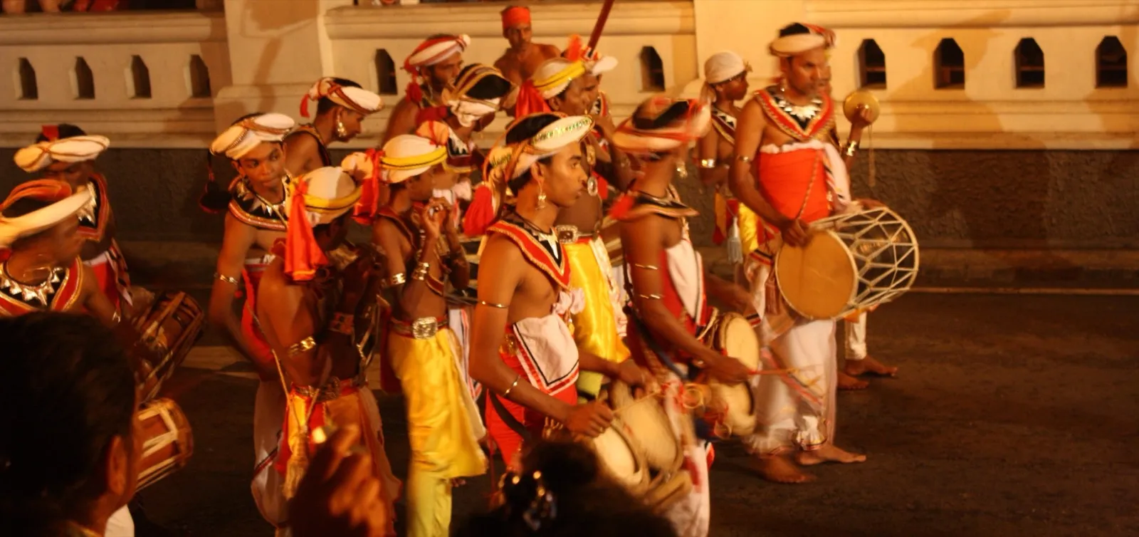 Drummers and dancers in red, white and gold costume parading at night in Kandy's Esala Perahera, a flaming torch behind them