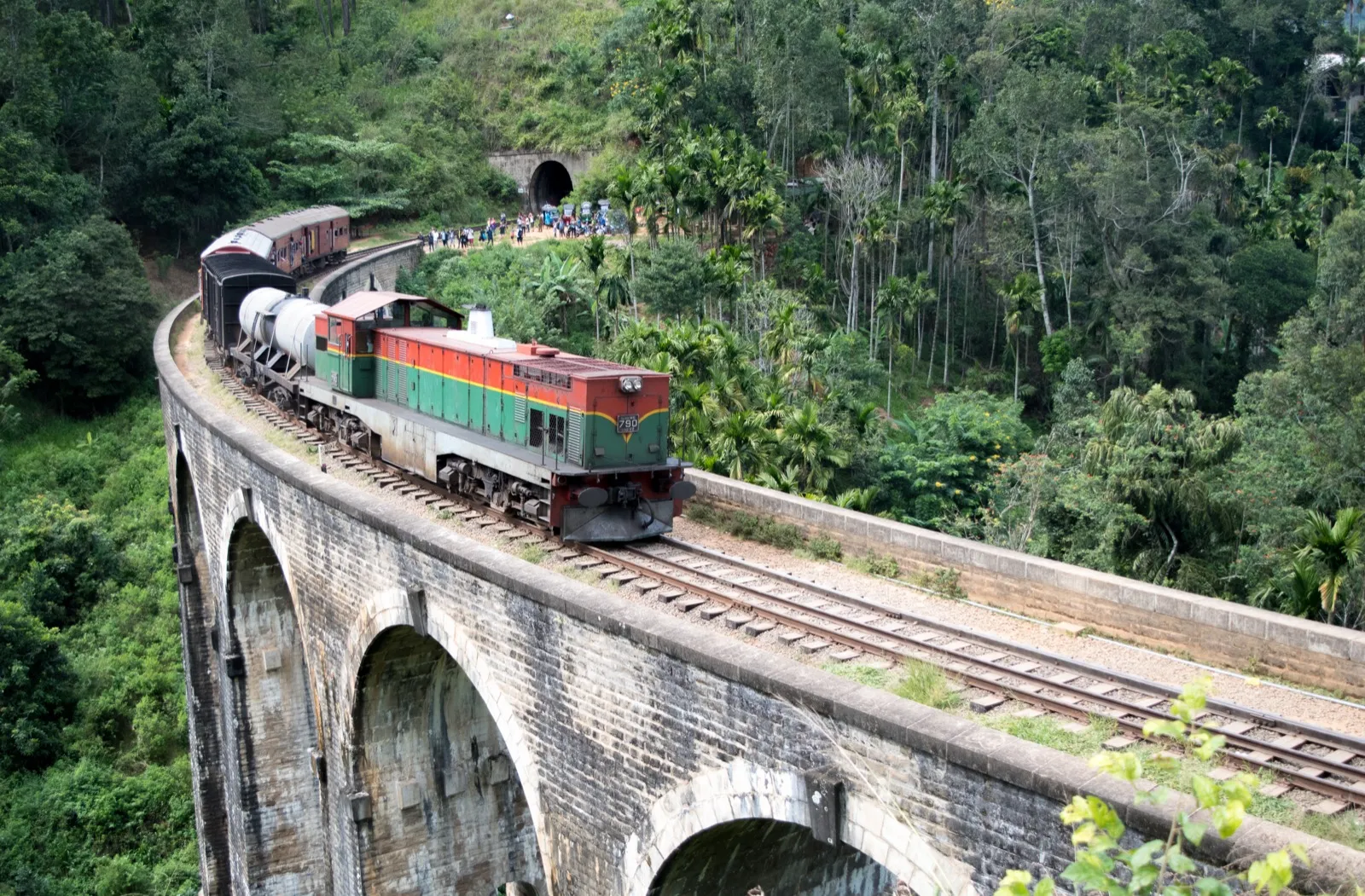 A green and red diesel train crosses the stone arches of the Nine Arch Bridge, surrounded by jungle