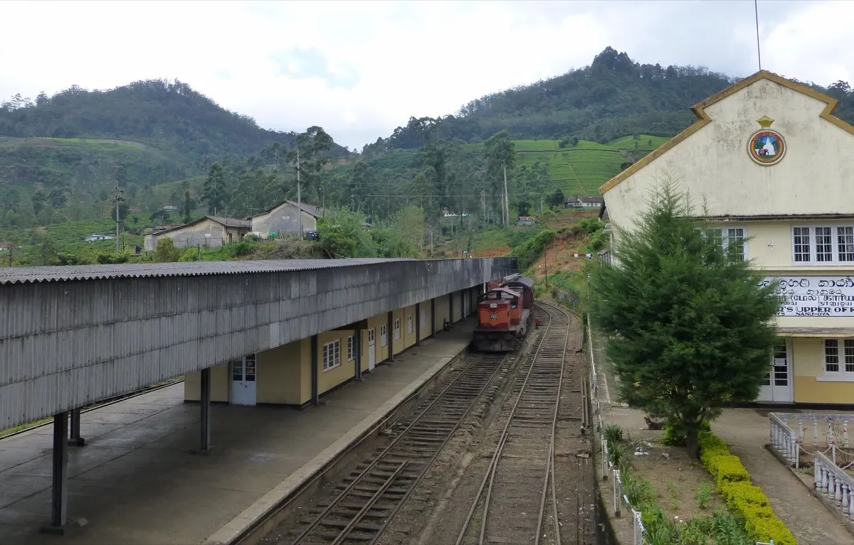 A train waiting at Nanu Oya railway station below tea-covered hills, the long platform roof on the left and the station building on the right