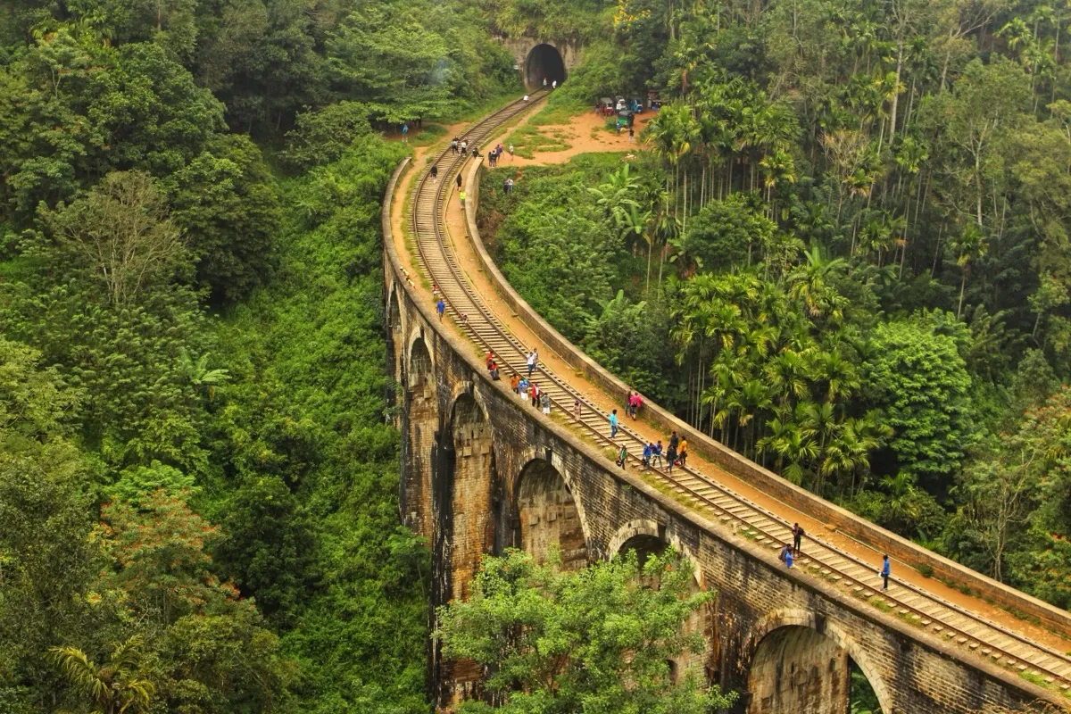 Visitors walking along the railway tracks across the Nine Arch Bridge, between jungle and areca palms