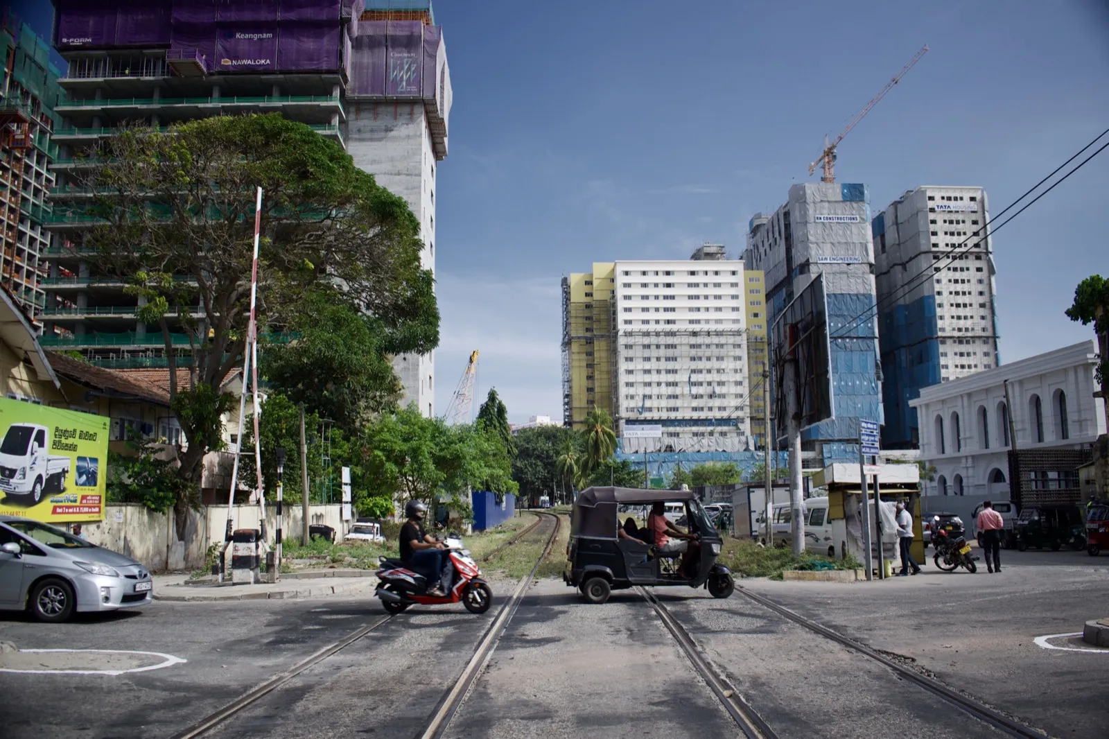 A three-wheeler tuk-tuk and a scooter crossing railway tracks on a Colombo street, with tall buildings behind