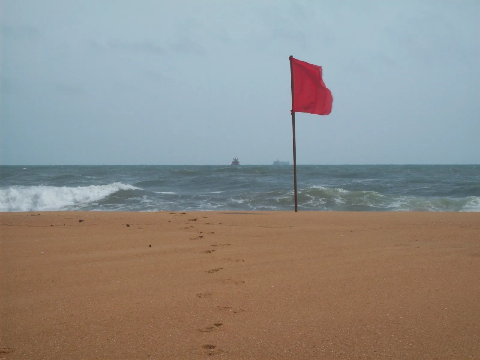 A red flag on a pole at Mount Lavinia beach, with rough surf breaking on the shore under an overcast sky
