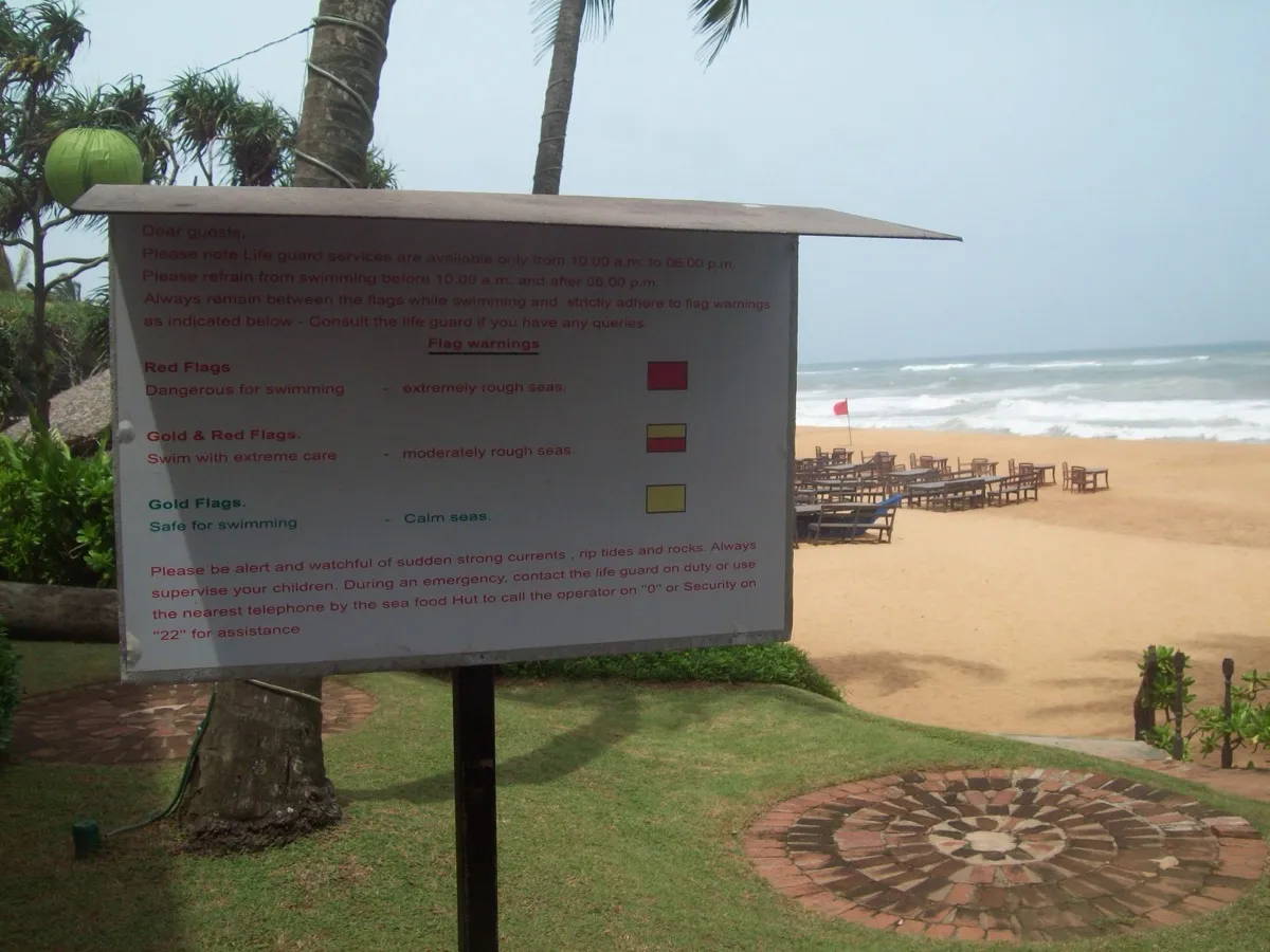 A printed beach-safety notice explaining red, red-and-gold and gold warning flags, with sunbeds, rough surf and a red flag on Mount Lavinia beach behind it