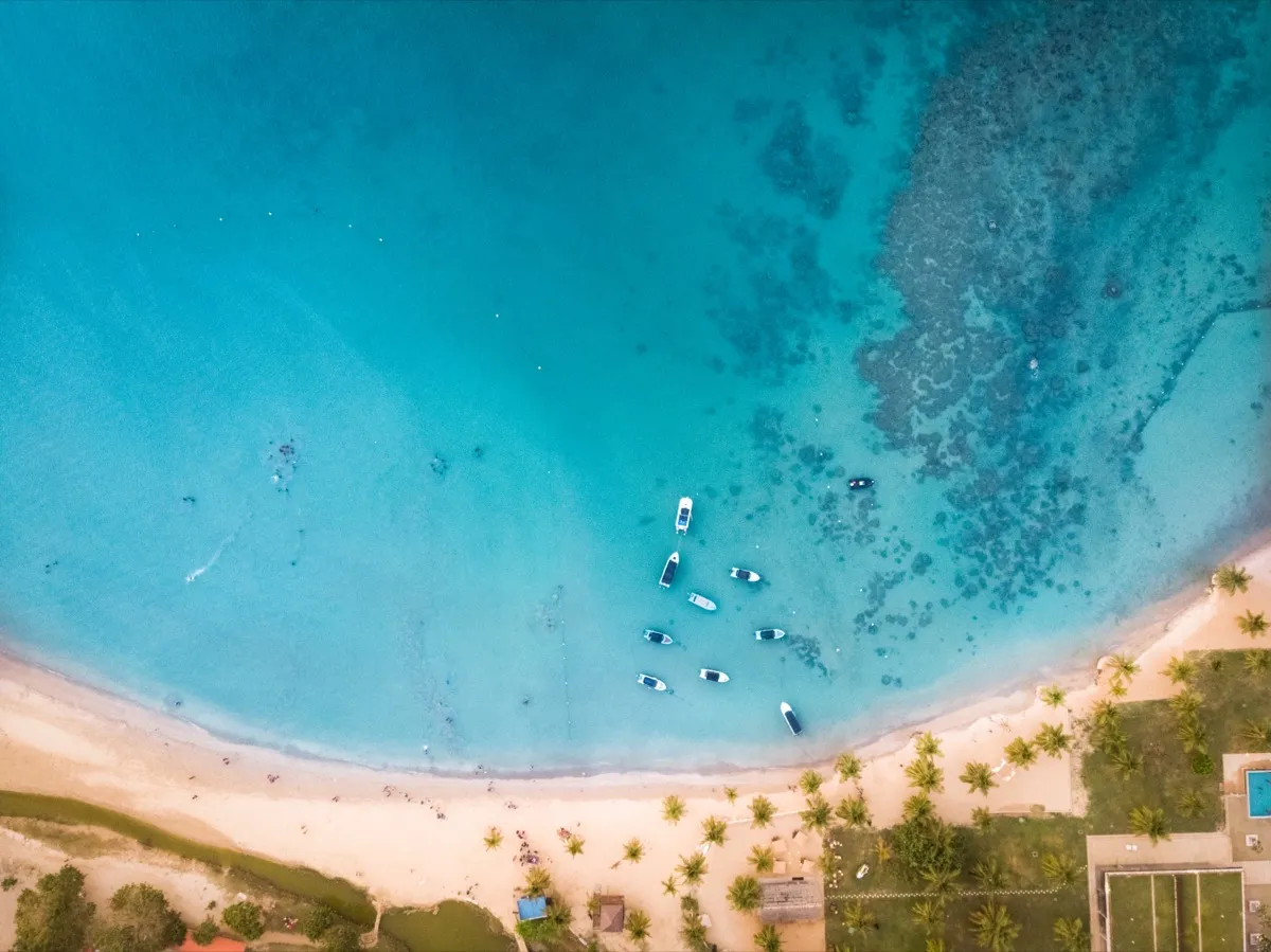 An aerial view of the curving beach and shallow turquoise bay at Pasikudah, with small boats moored offshore and a reef showing through the water