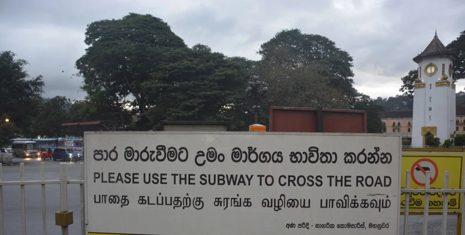A roadside sign in Kandy reading 'Please use the subway to cross the road' in Sinhala, English and Tamil, with a white clock tower behind