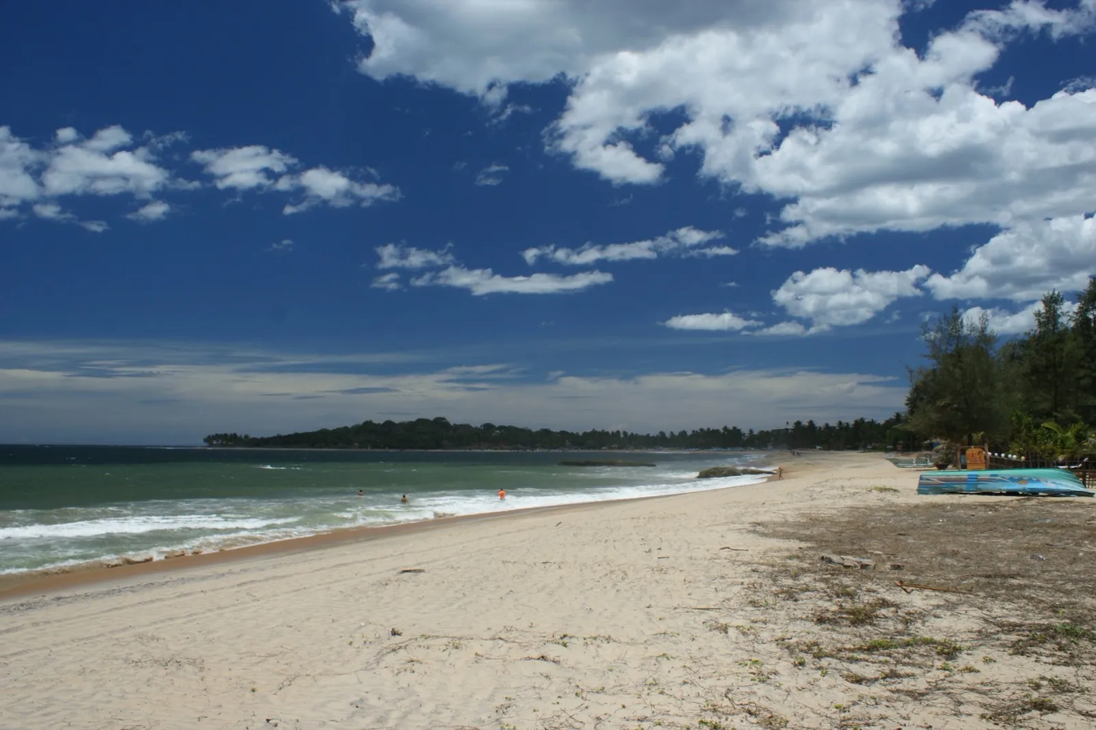 The wide sandy beach at Arugam Bay curving south to a wooded headland, small waves breaking under a deep blue sky with white clouds