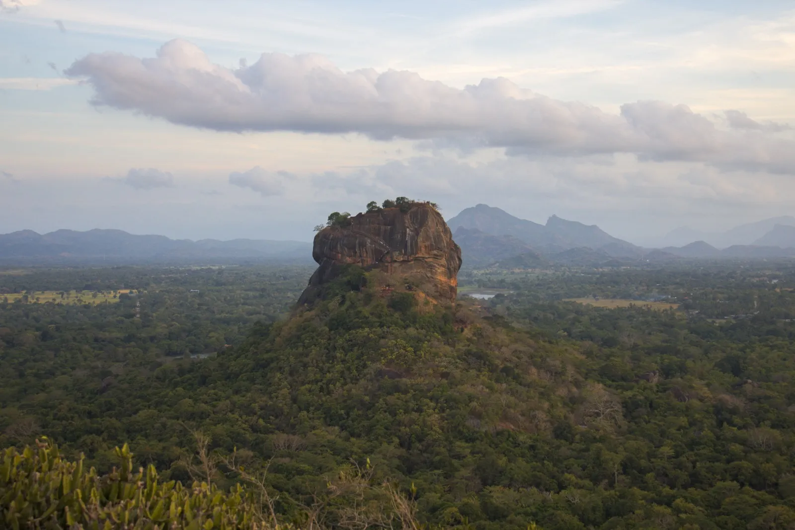 Sigiriya, a huge flat-topped rock, rises out of green forest with hills behind it under a cloudy sky