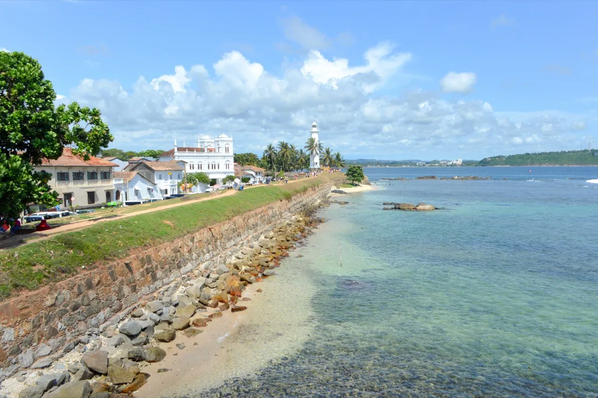 A grassy path along the old stone sea wall of Galle Fort leads to a white lighthouse beside clear blue water