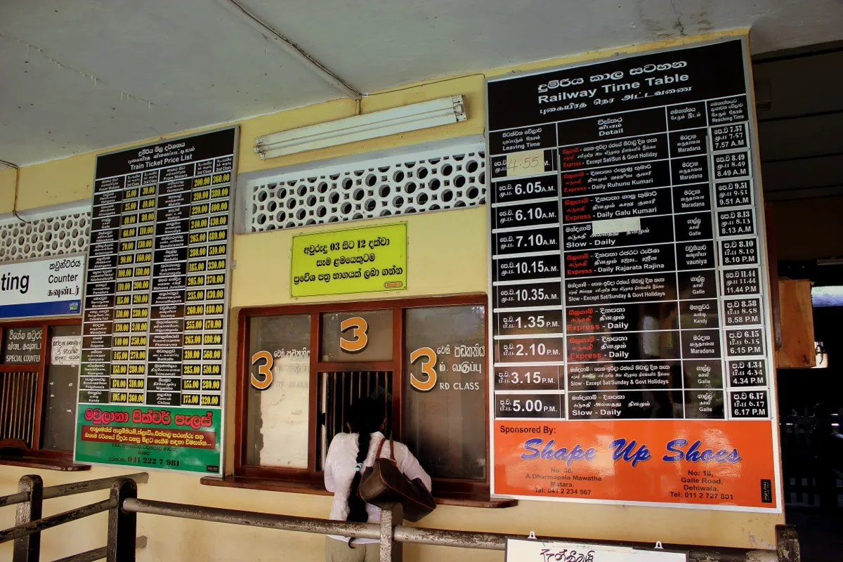 A railway ticket counter marked 3rd class, between a printed ticket price list and a train timetable board
