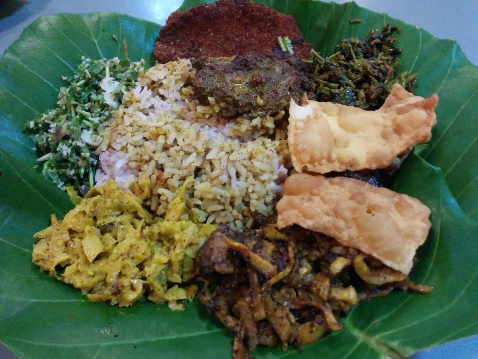 A Sri Lankan rice and curry plate on a green leaf, with rice surrounded by vegetable curries, greens and crisp fritters