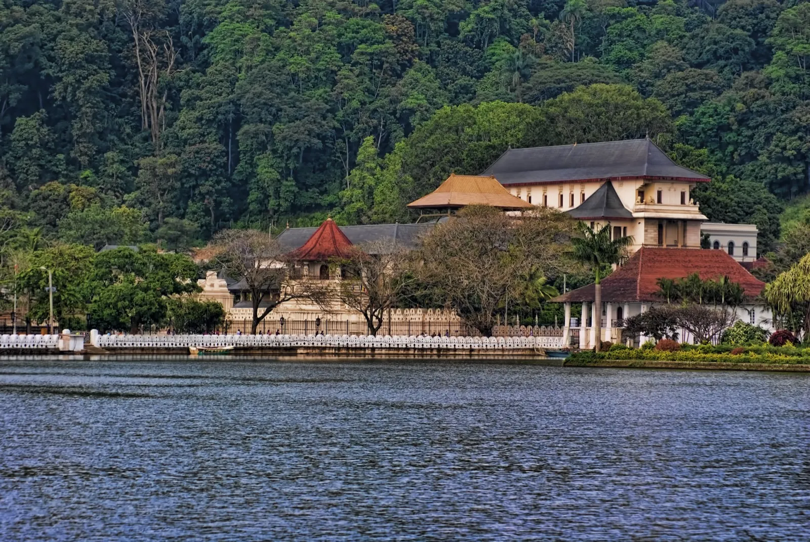 The Temple of the Tooth in Kandy, white walls and red-roofed buildings beside the lake below a forested hill