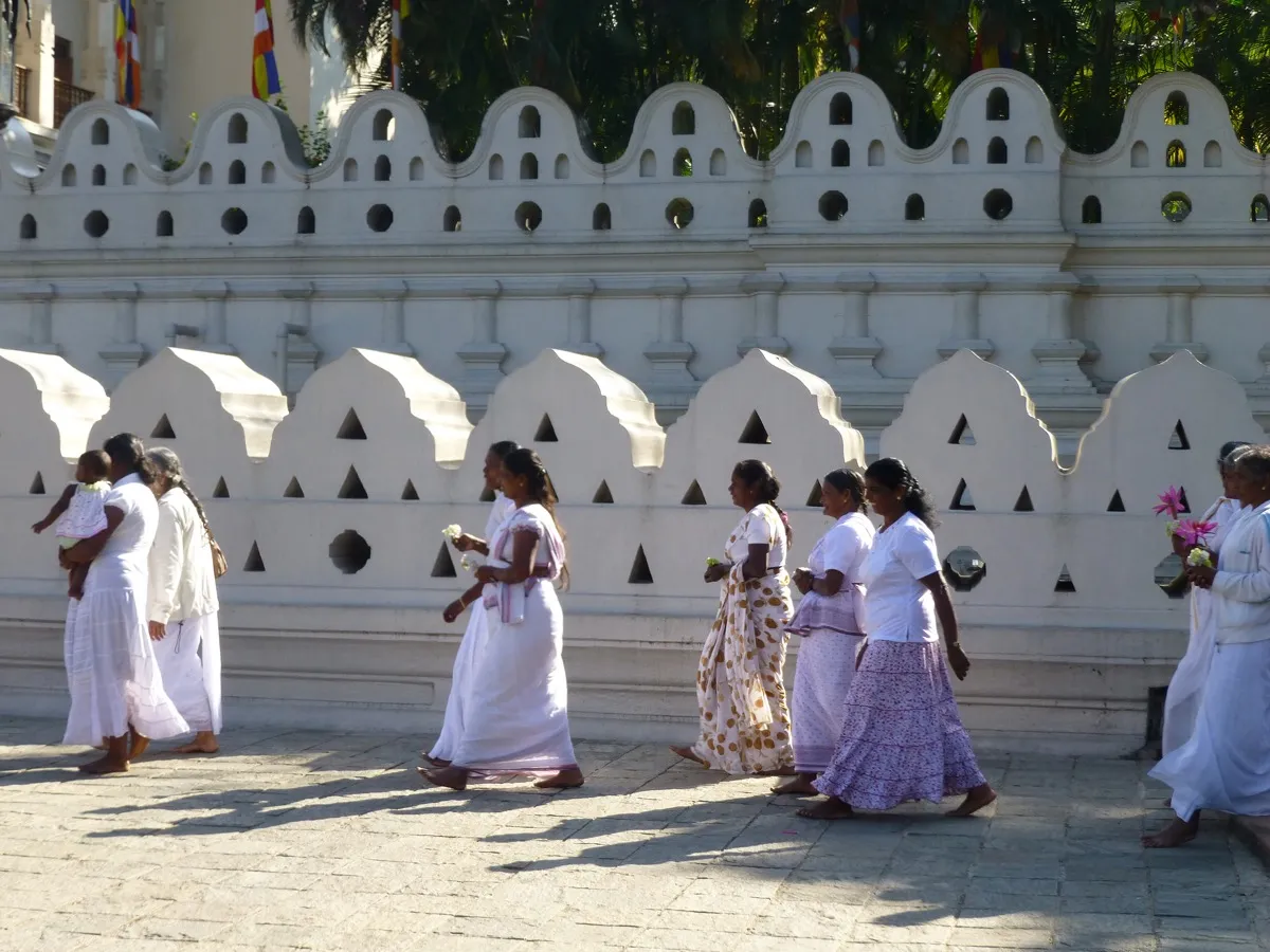 Women in white clothes and long skirts walk barefoot beside the white wall of the Temple of the Tooth, carrying flowers