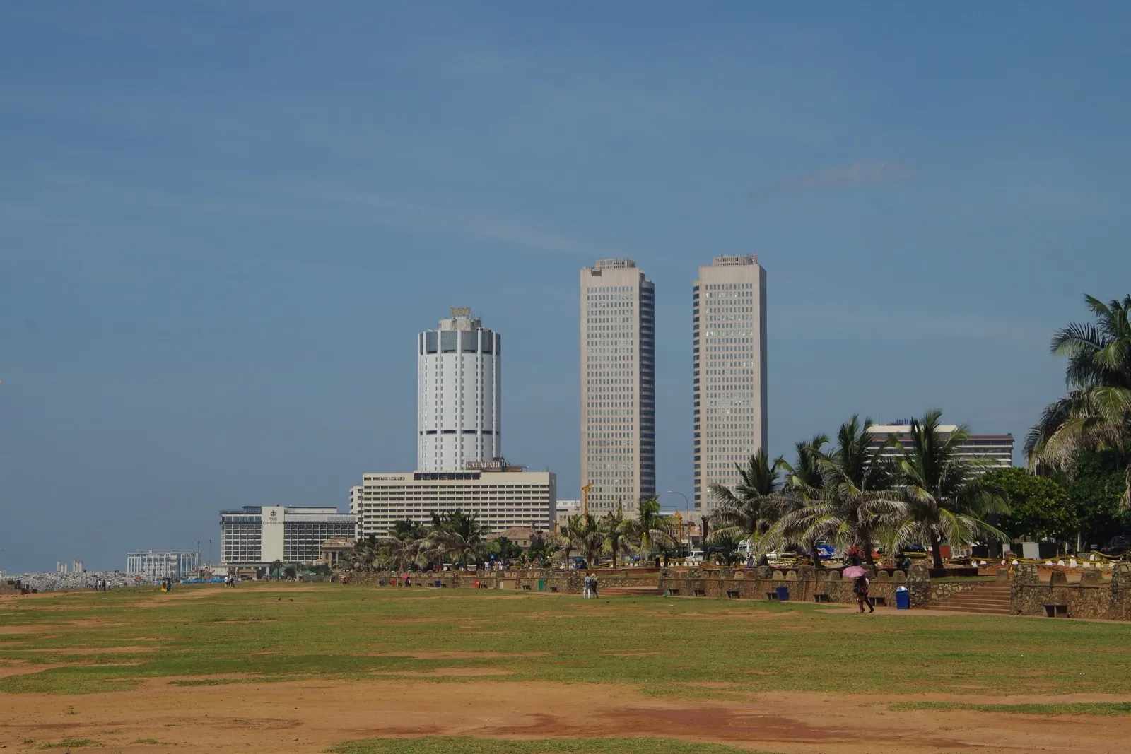 A wide grassy seafront lawn in Colombo with a few people walking, palm trees, and tall office towers rising behind under a blue sky