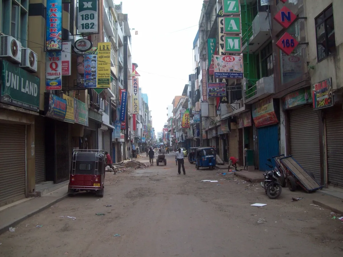 A narrow city street lined with closed shop shutters and dozens of colourful hanging shop signs, with a tuk-tuk, a motorbike, handcarts and a few people in the road