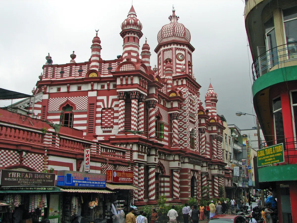 A large mosque painted in red and white stripes and checks, with onion domes and a clock tower, rising above small shops and a busy street