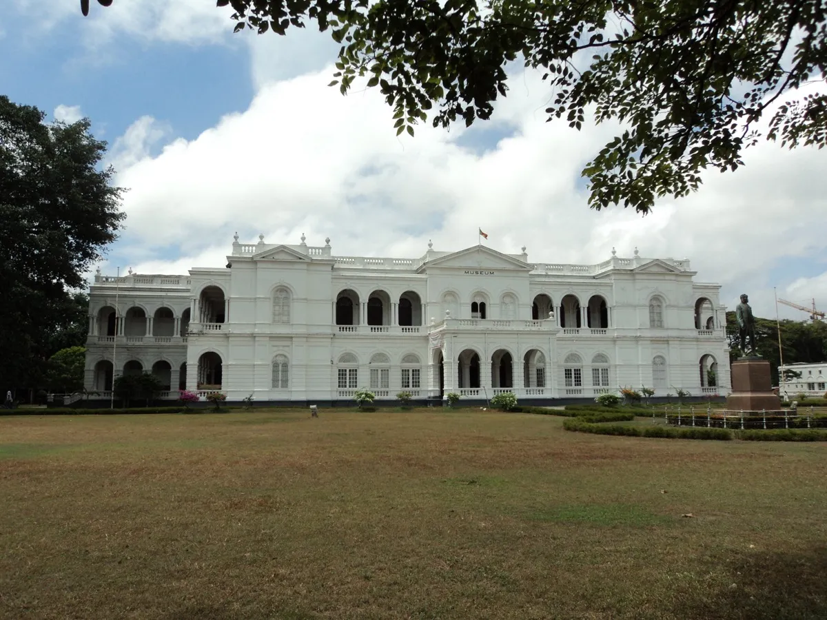 A long white two-storey colonial building with arched verandas behind a wide lawn, with a bronze statue of a man in nineteenth-century dress on a plinth at right