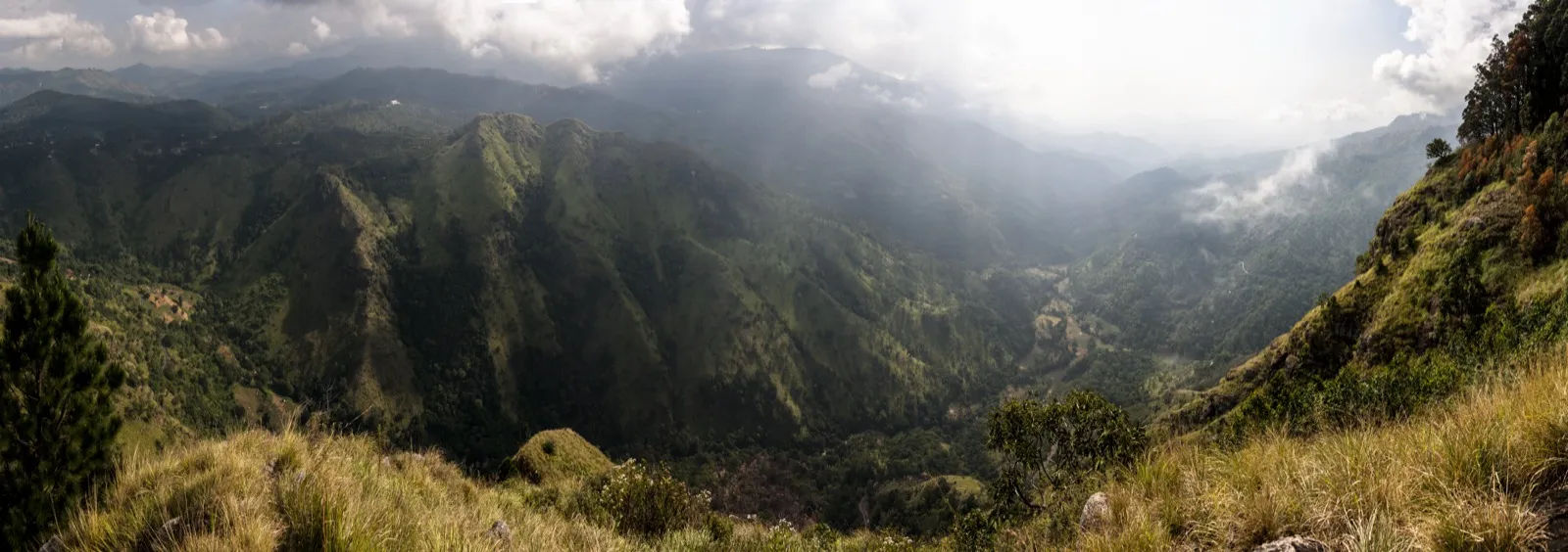 Wide panorama of the Ella Gap, a deep green valley between steep grassy ridges opening toward the hazy lowlands of Sri Lanka