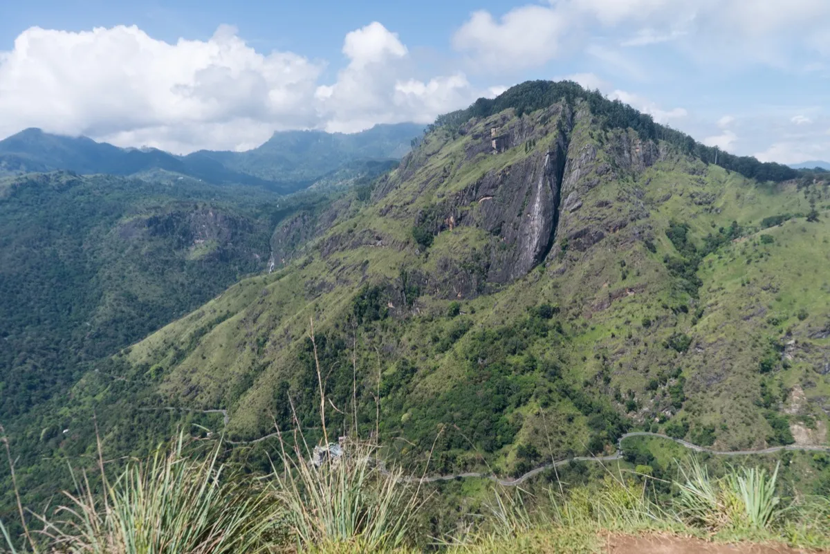 Ella Rock's sheer grey cliff and green slopes seen across the valley from Little Adam's Peak, a road winding along its foot