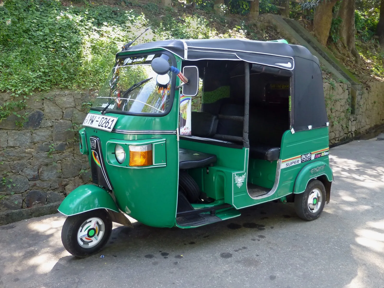 A green Sri Lankan tuk-tuk with a black canvas roof parked on a quiet road beside a stone wall