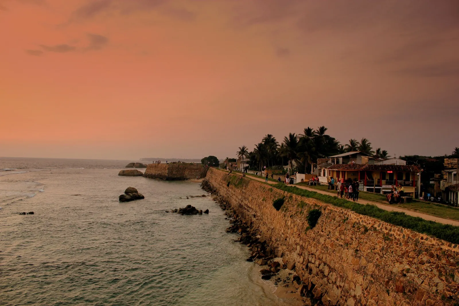 The old stone sea wall of Galle Fort at sunset, with people strolling along the path on top and houses beside it